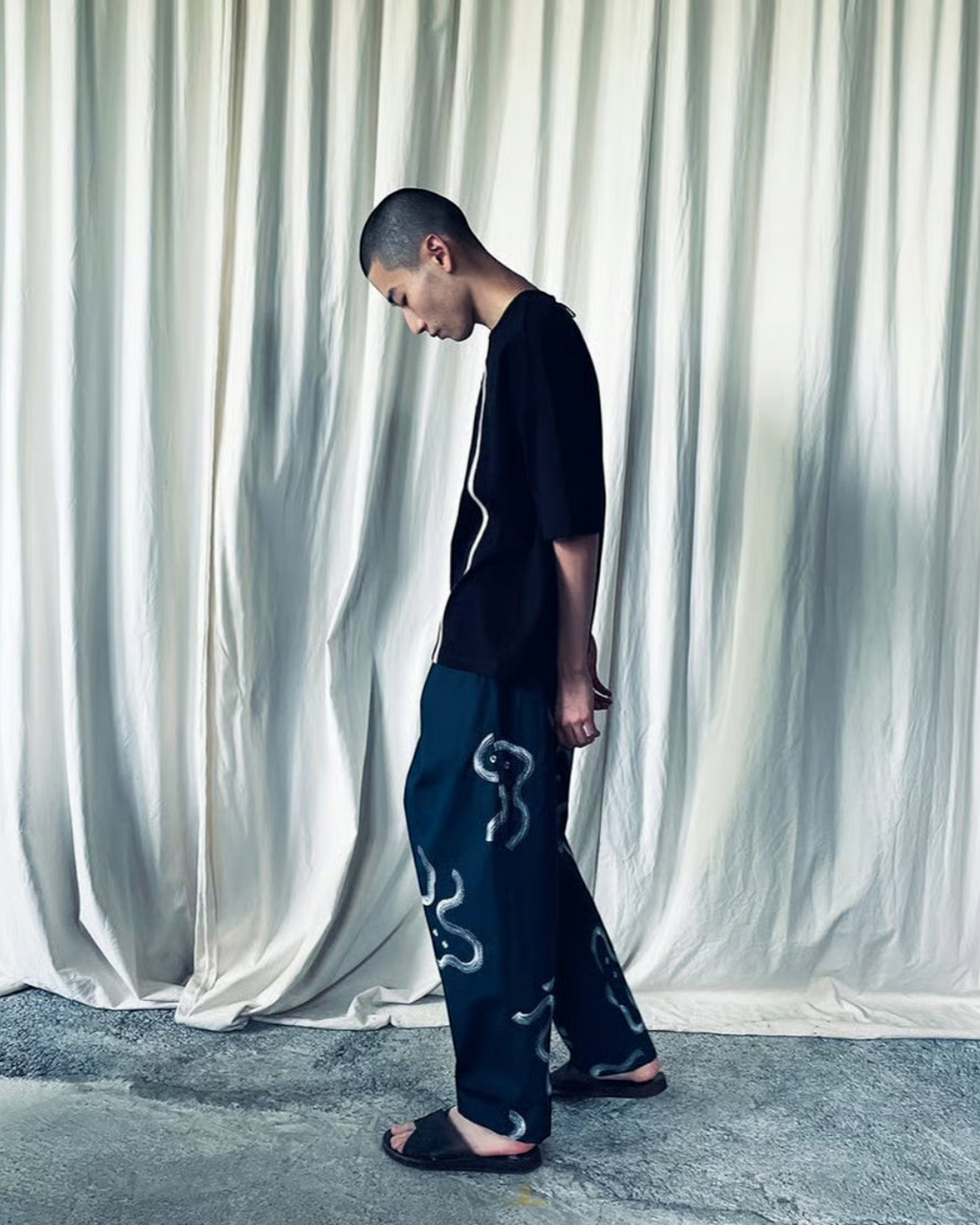 CALCÈLの黒いTシャツを着用し、お地蔵さんの刺繍が施されたパンツを合わせた男性モデルの全身ショット