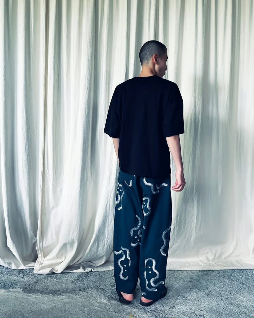 CALCÈLのohta黒色コットンTシャツを着用し、お地蔵さんのシルエット柄パンツを合わせた男性の背面ショット