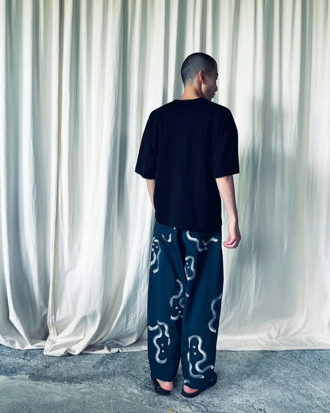CALCÈLのohta黒色コットンTシャツを着用し、お地蔵さんのシルエット柄パンツを合わせた男性の背面ショット