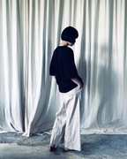 CALCÈLのohta黒色コットンTシャツを着用し、白いカーテンを背景に横向きで立つモデル