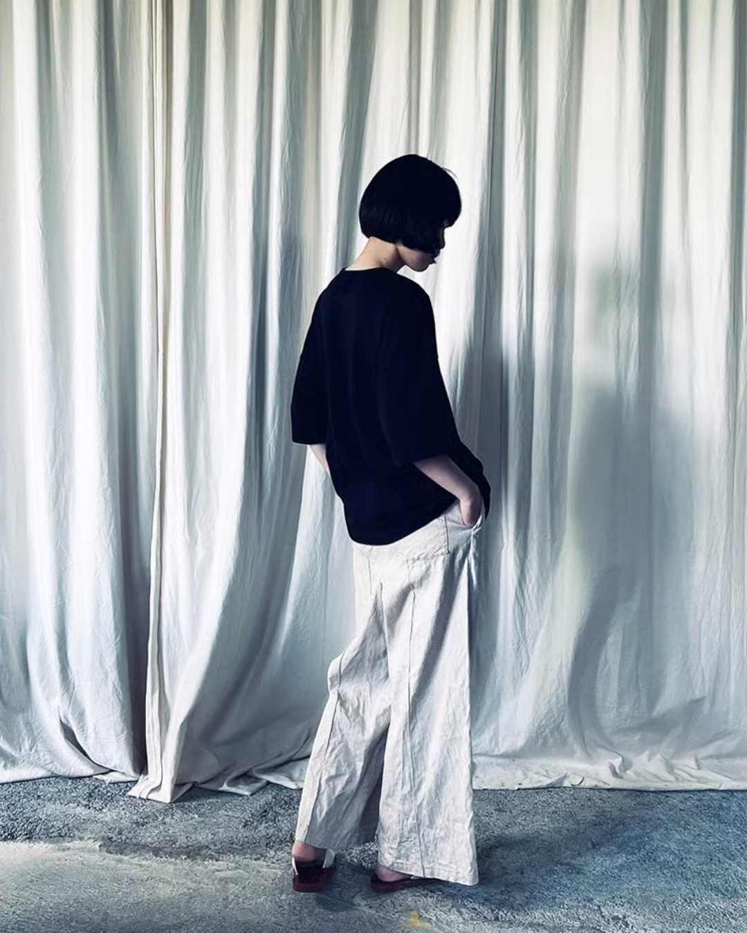 CALCÈLのohta黒色コットンTシャツを着用し、白いカーテンを背景に横向きで立つモデル